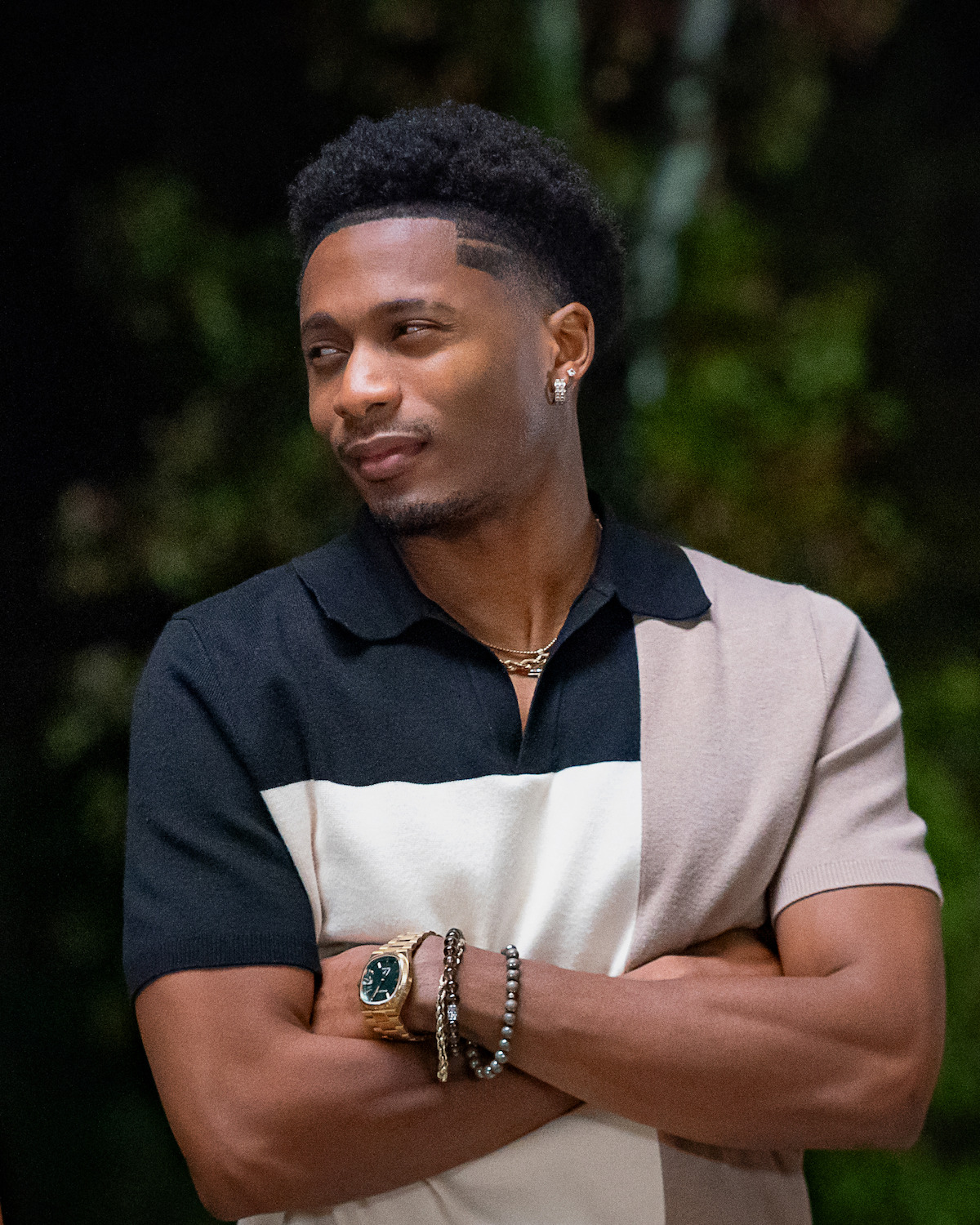 A stylish man stands outdoors at night with his arms crossed, wearing a color-block polo shirt, gold jewelry, and a watch, with a confident expression and blurred greenery in the background.