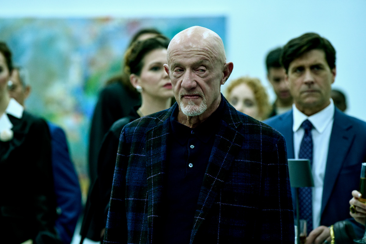 A serious bald man in a plaid jacket stands among formally dressed people at a social gathering or art event, with a soft focus background and a subdued, contemplative mood.