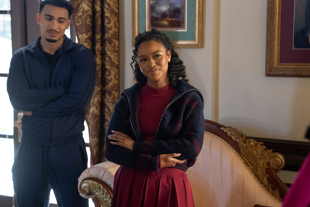 Xavier Smalls as Angel and Bailey Tippen as Sylvie stand indoors in a warmly lit, elegant room with ornate furniture and framed art. The woman in front is smiling with arms crossed, while the man behind her stands with folded arms near a window.