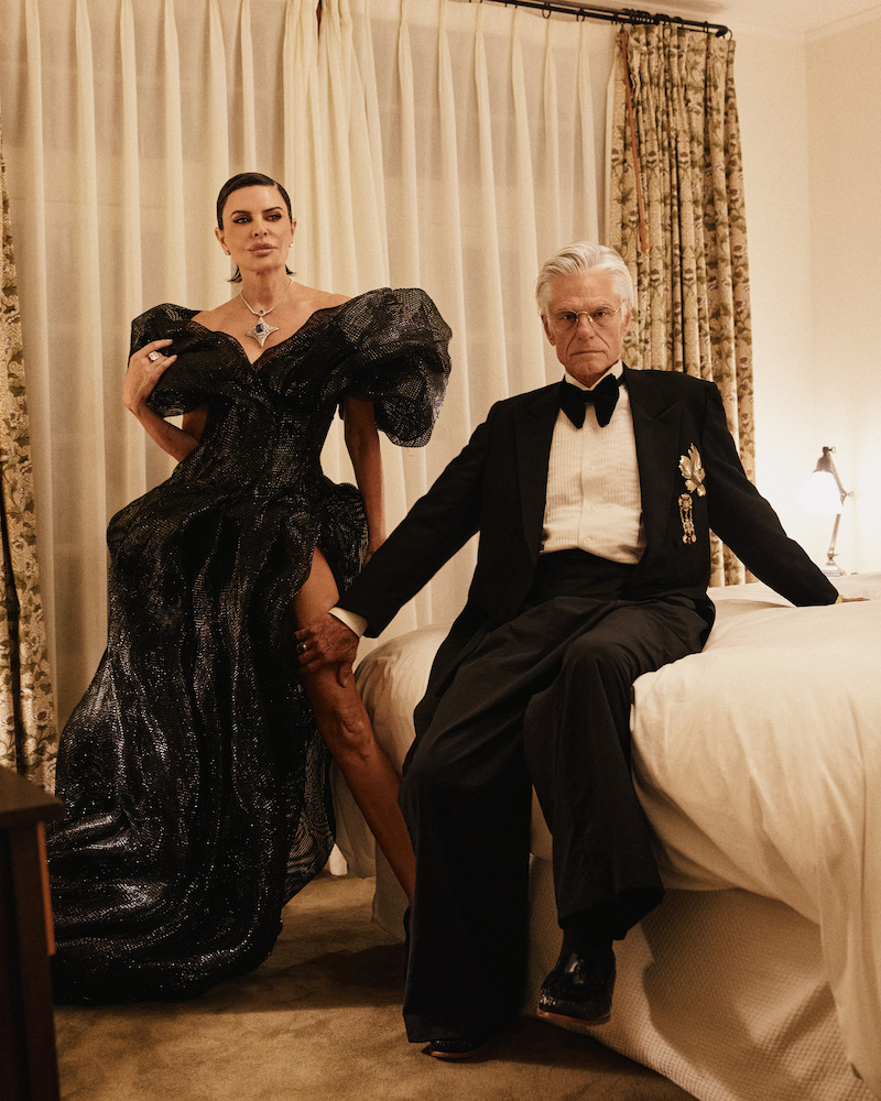 A woman in a dramatic black gown stands confidently beside a man in a tuxedo sitting on a bed in an elegantly lit bedroom with classic curtains and warm, neutral decor.