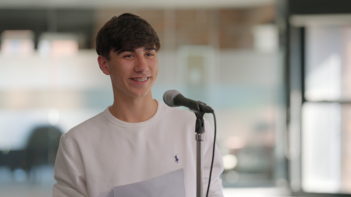 Teen boy smiling, standing at a microphone with papers in hand, in a bright, modern indoor setting. The mood is confident and relaxed, suggesting he’s giving a speech or presentation.