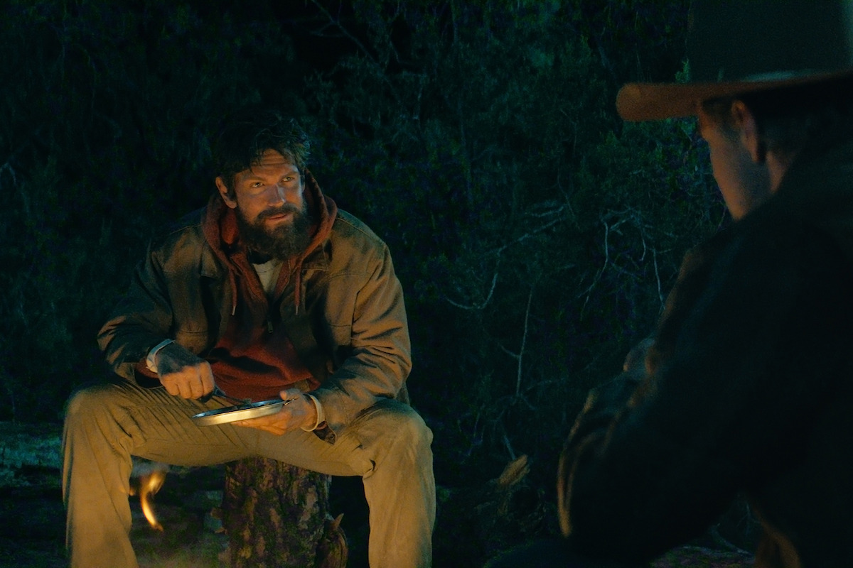 Two men sit around a campfire at night in the woods, one holding a plate and eating utensil, both dressed in rugged outdoor clothing, illuminated by firelight.