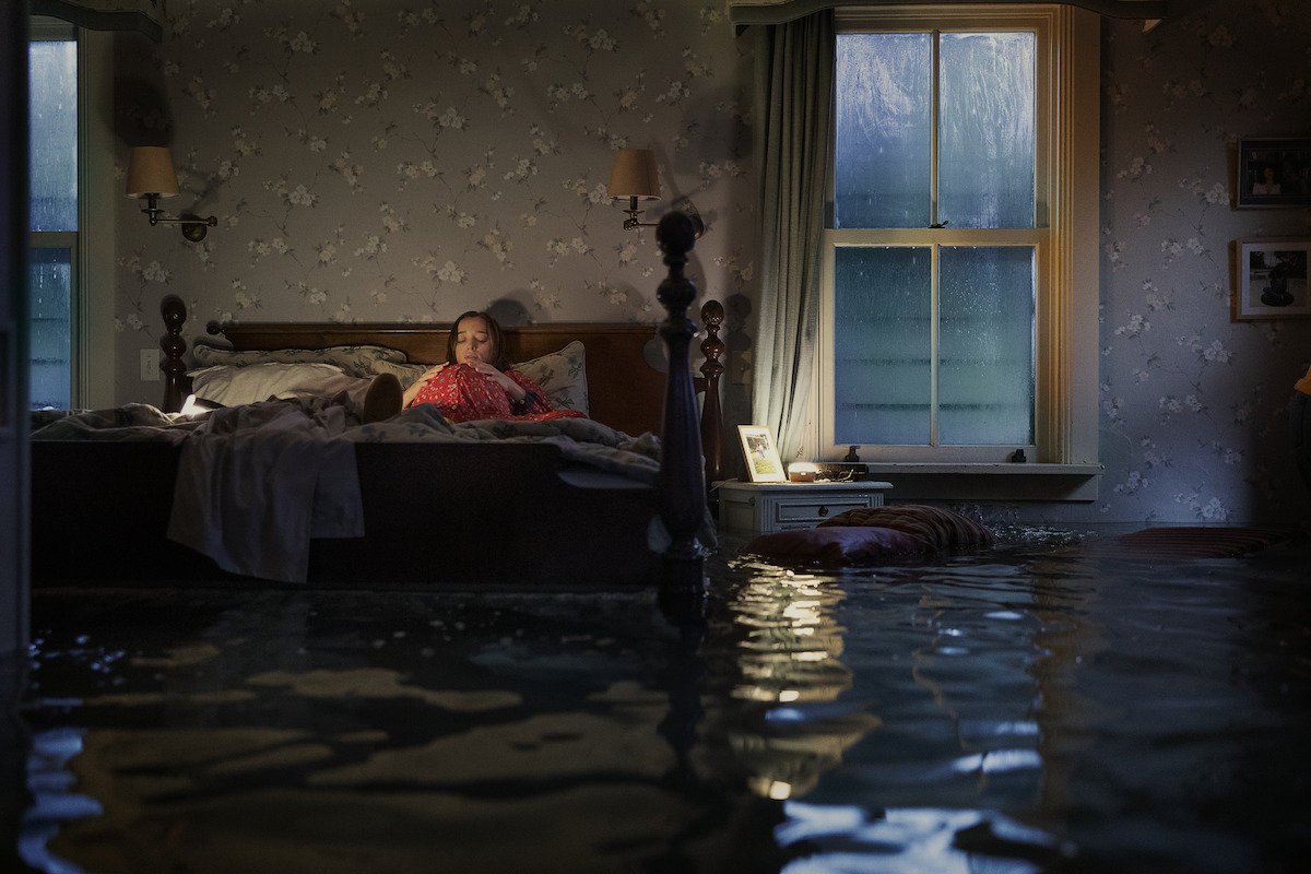 A woman lies on a bed in a dimly lit bedroom with patterned wallpaper, as floodwater covers the floor, reaching the furniture and reflecting light from a window and lamps, creating a dramatic and surreal atmosphere.