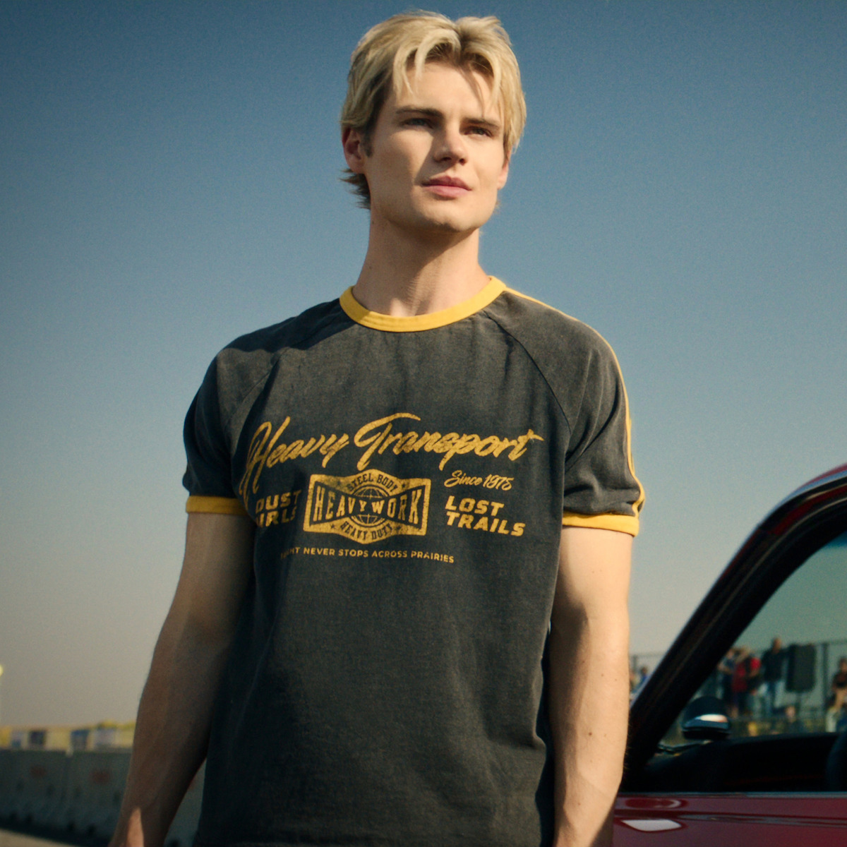 Young man with blond hair stands outdoors beside a red car, wearing a dark t-shirt with yellow trim and text, under a clear blue sky, with a parking lot and buildings in the background.
