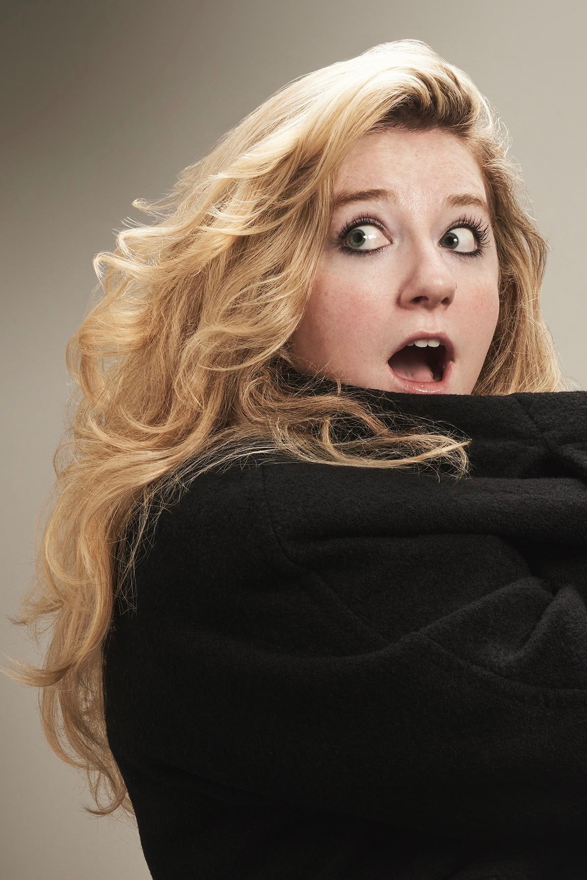 Young woman with long blonde hair in a black coat looking surprised or scared, turning her head over her shoulder in a studio with neutral background.