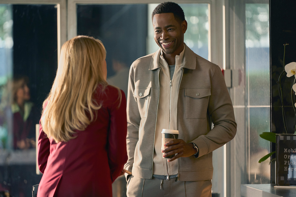 Man smiling and holding a coffee cup while talking to a woman with long blonde hair in an indoor cafe or office setting, with large windows and a modern, casual atmosphere.