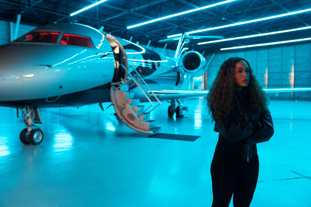 Uma mulher de cabelos cacheados posa com confiança em um hangar de aviões moderno e bem iluminado, ao lado de um elegante jato particular. A luz azul refletida no piso brilhante cria uma atmosfera futurista.