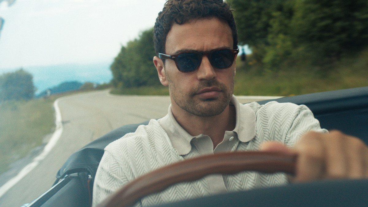 Man wearing sunglasses driving a convertible car on a winding road with trees and greenery on both sides, appearing focused and serious in a relaxed, outdoor rural setting.
