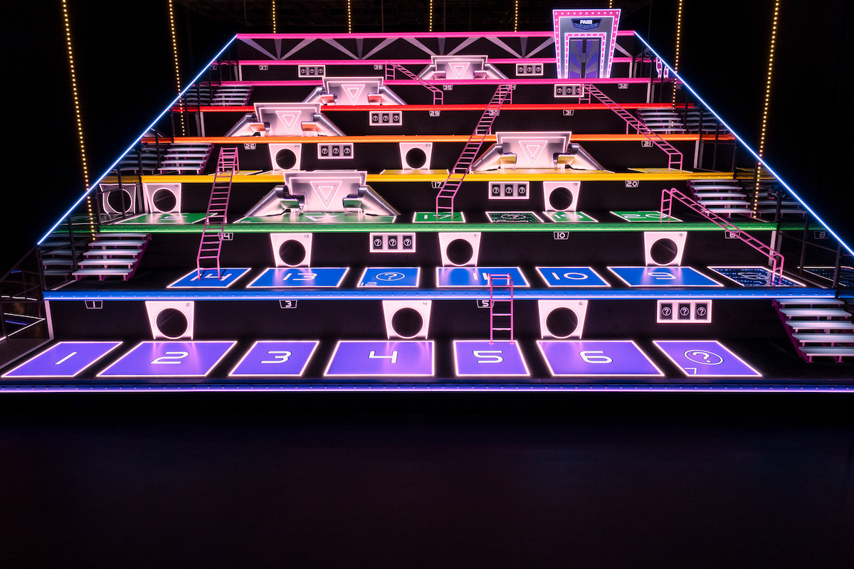 A brightly lit, multilevel game show set with numbered sections, colorful lights, ladders, and platforms. The environment is vibrant and energetic, suggesting a playful, competitive mood.