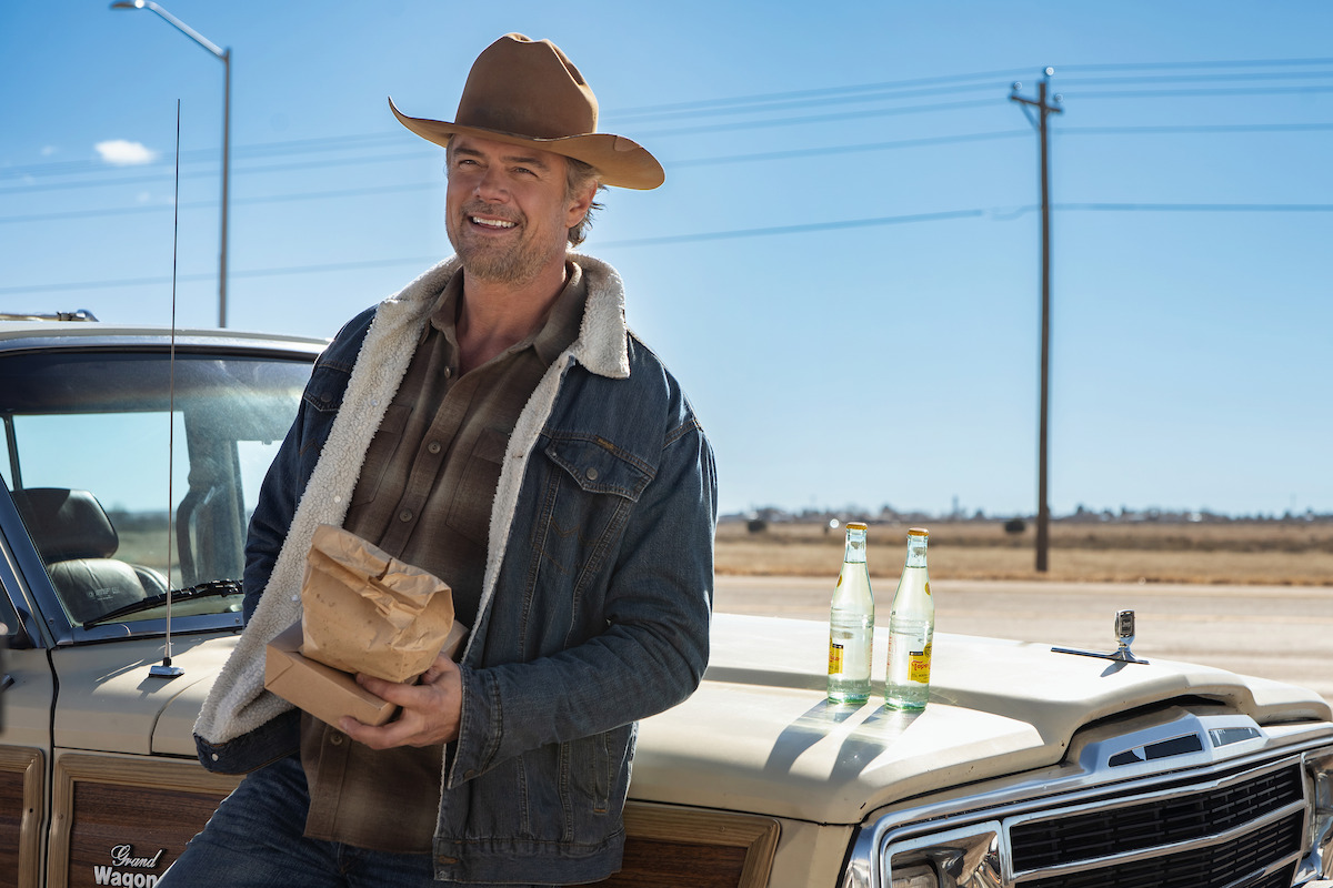 Man in cowboy hat and denim jacket stands by vintage SUV holding paper bag on a sunny rural roadside. Two glass soda bottles sit on the car hood, with open field and power lines in the background.