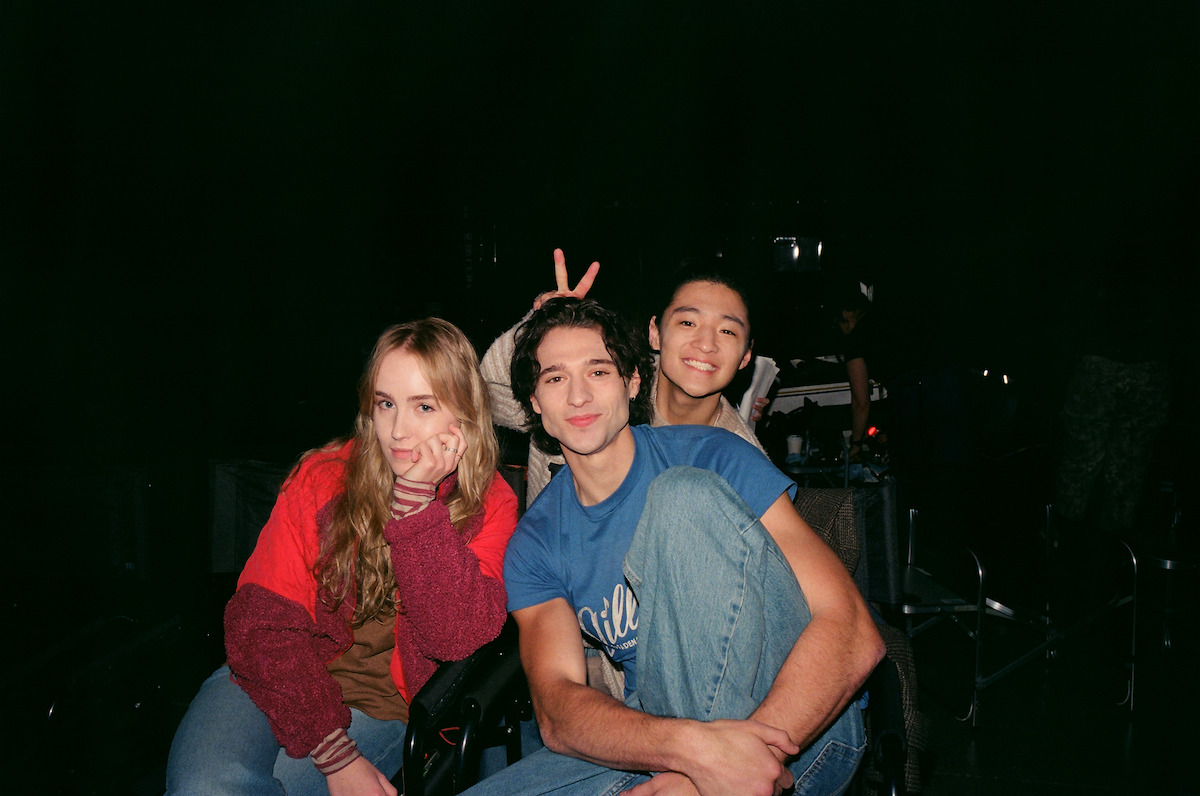 Three young adults sit close together in a dimly lit room, posing casually. The woman on the left has long blonde hair and a red jacket, while the two men on the right smile, with one making bunny ears behind the other.