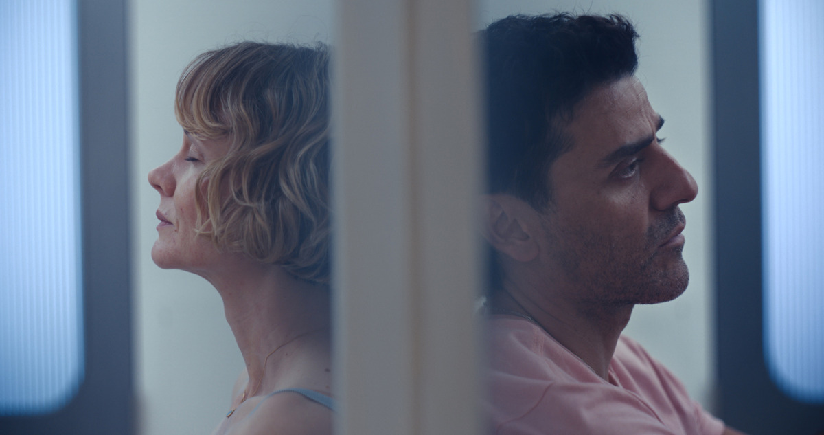 A woman and a man sit back-to-back separated by a wall in a modern, softly lit room, both appearing contemplative or distant, with subtle blue lighting in the background.