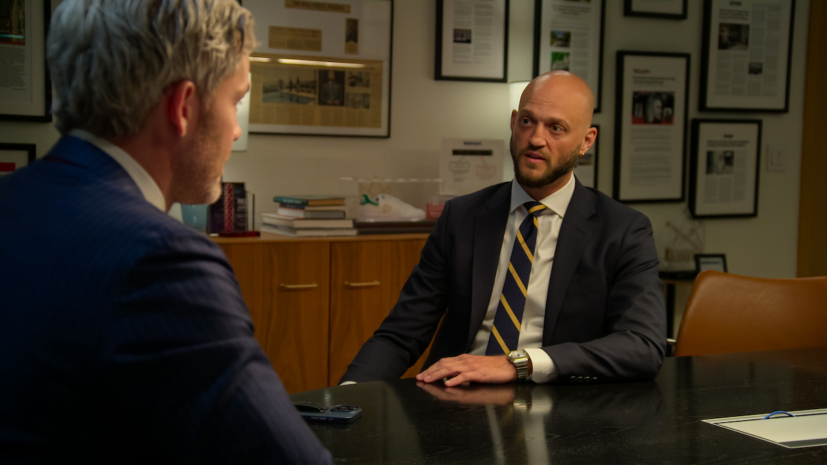 Two men in suits have a serious discussion in a modern office with framed articles on the wall, books, and documents on a cabinet, suggesting a professional and formal atmosphere.