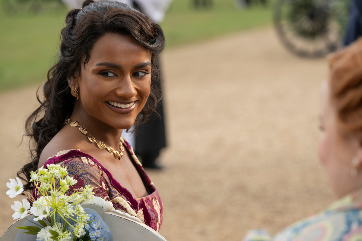 Woman in elegant dress smiling outdoors on a pathway, with blurred people and greenery in the background, suggesting a historical or period setting.