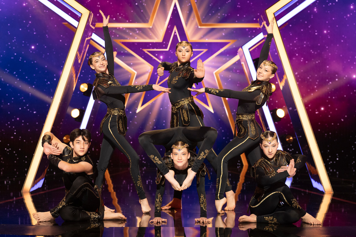 K-STAR in black and gold costumes pose dynamically on a stage with a vibrant purple star-lit backdrop and bright lights, suggesting a talent or dance competition setting.