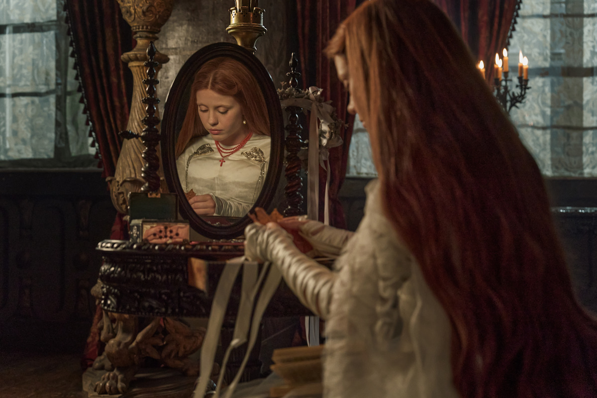 A woman in a medieval-style dress sits at an ornate vanity, braiding her hair, with her reflection visible in a mirror. The dimly lit, richly decorated room adds an atmosphere of quiet introspection.