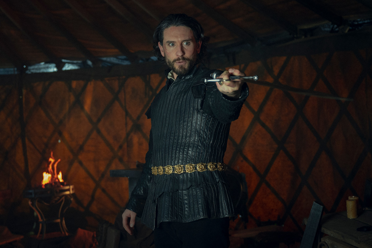 A man in medieval-style dark leather armor stands in a dimly lit tent, holding a sword out with a serious expression. The environment is rustic, with warm firelight casting shadows, creating a tense and dramatic mood.