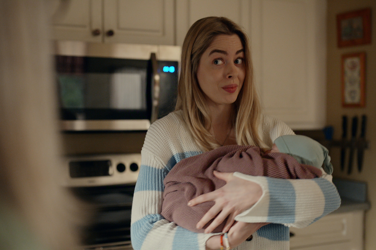 Woman in a kitchen holding a baby wrapped in a blanket, wearing a striped sweater and making a facial expression, with cabinets, microwave, and utensils visible in the background. Warm indoor setting.