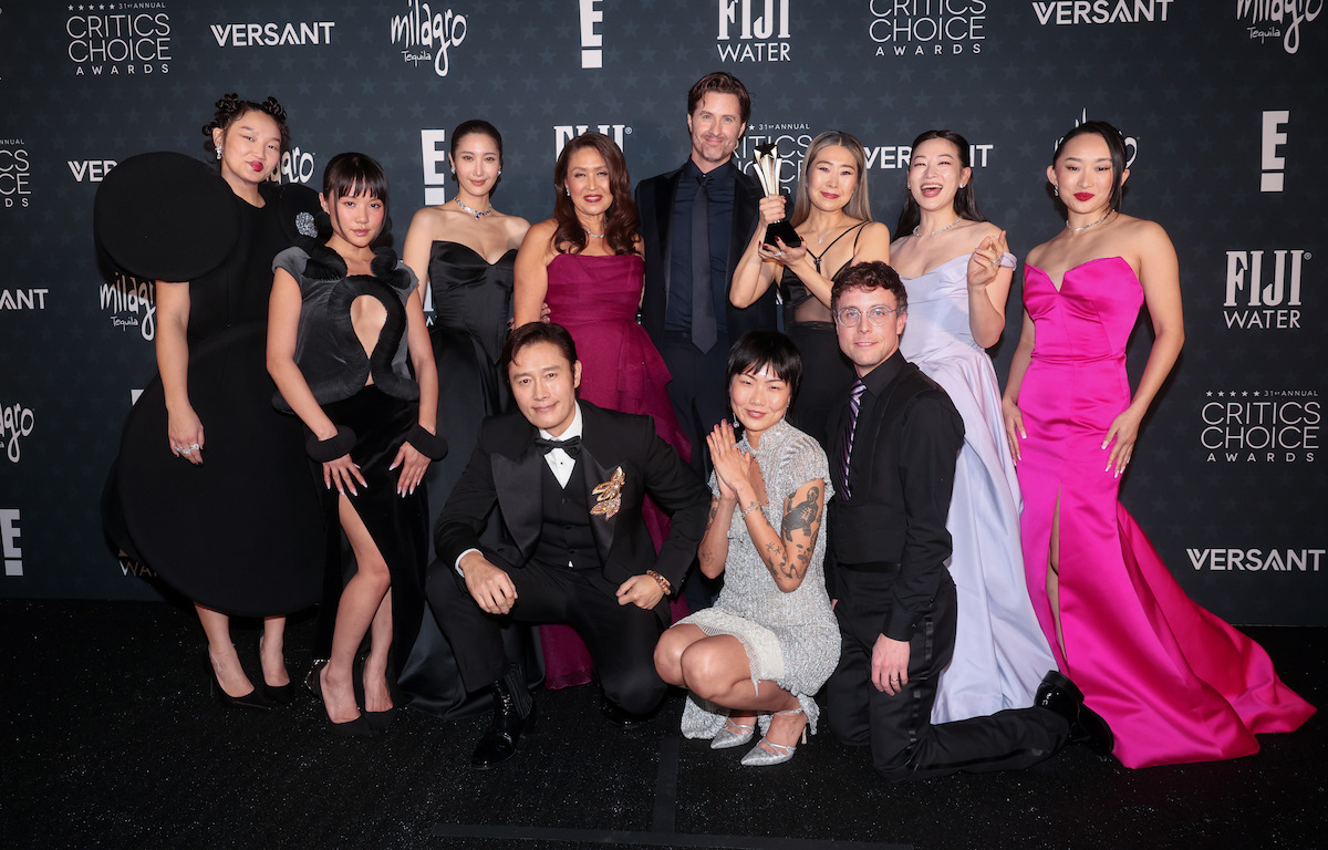 A group of elegantly dressed people pose together on a black carpet at an awards event with branded sponsor and event signage in the background.