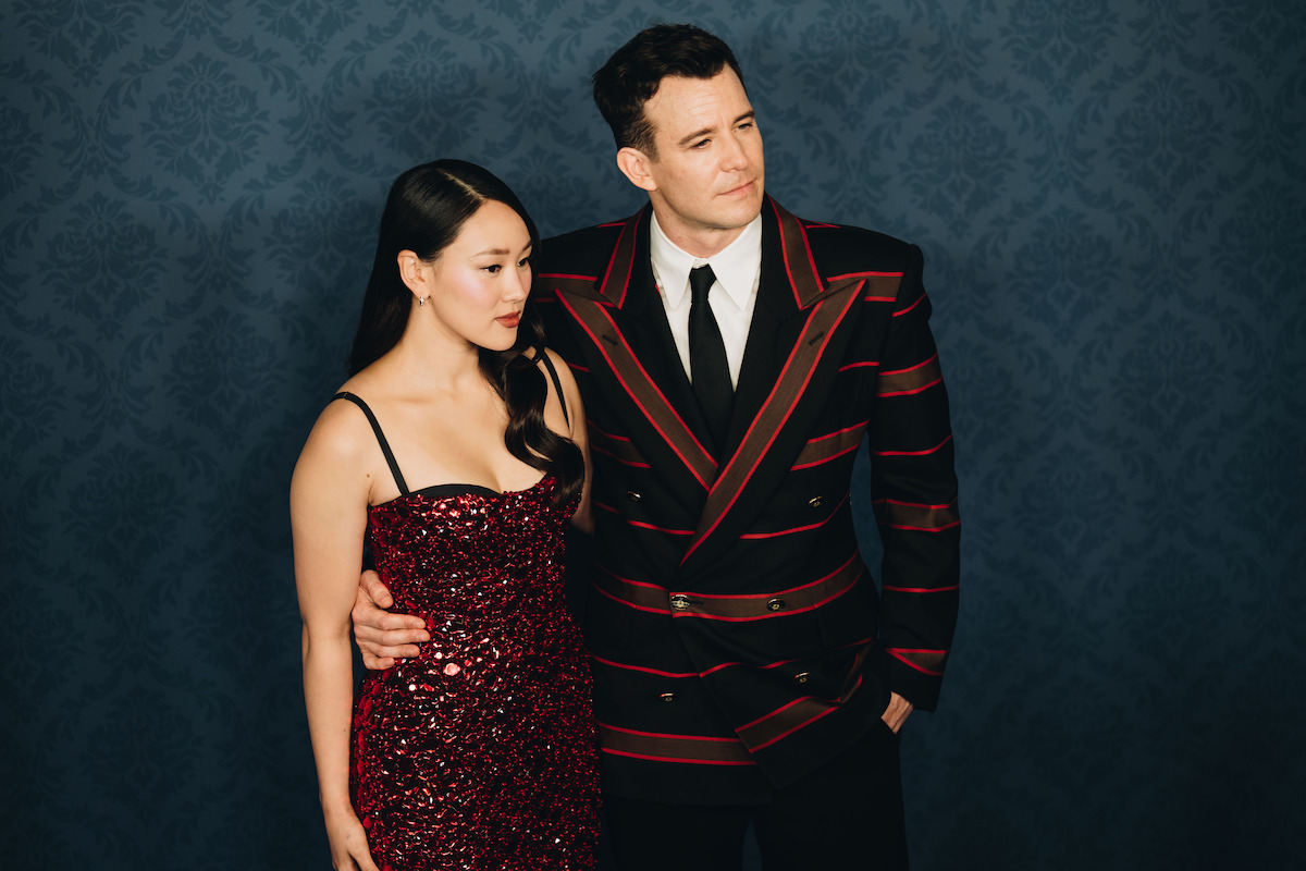 Mulher usando um vestido vermelho de paetês e homem de terno preto com listras vermelhas estão juntos diante de um painel azul estampado. É um evento formal.