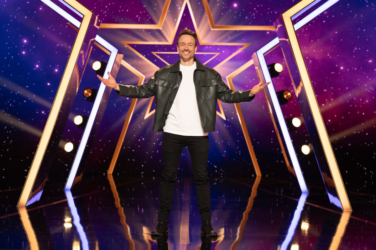 Jan Reinder wearing a black jacket stands smiling on a stage with bright lights, geometric gold lines, and a large star pattern and purple cosmic background, arms open wide in a welcoming gesture.