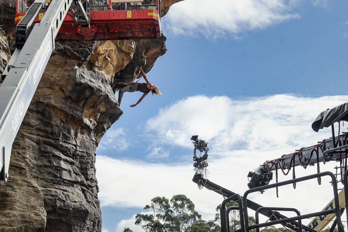 A person jumps off a rocky cliff face with safety equipment above, as camera cranes capture the action outdoors under a blue sky with clouds and trees in the background.