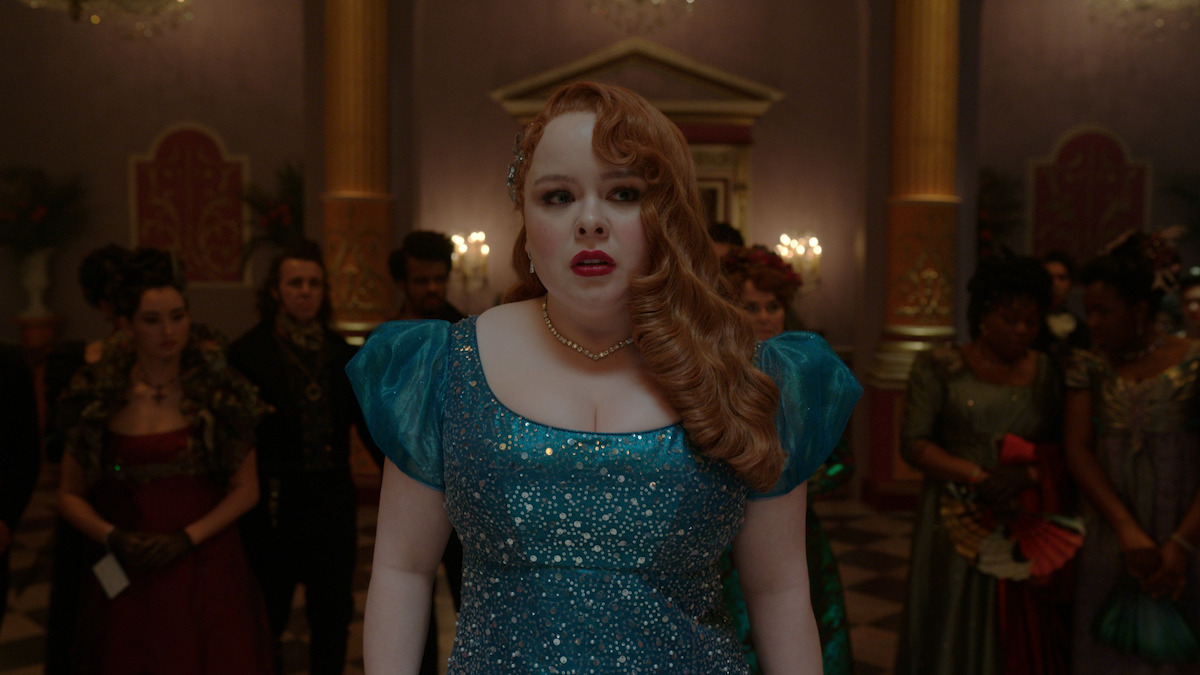 A woman in a sparkling blue gown stands in a grand, candlelit ballroom with ornate columns and people in period costumes behind her, creating a dramatic and elegant setting.