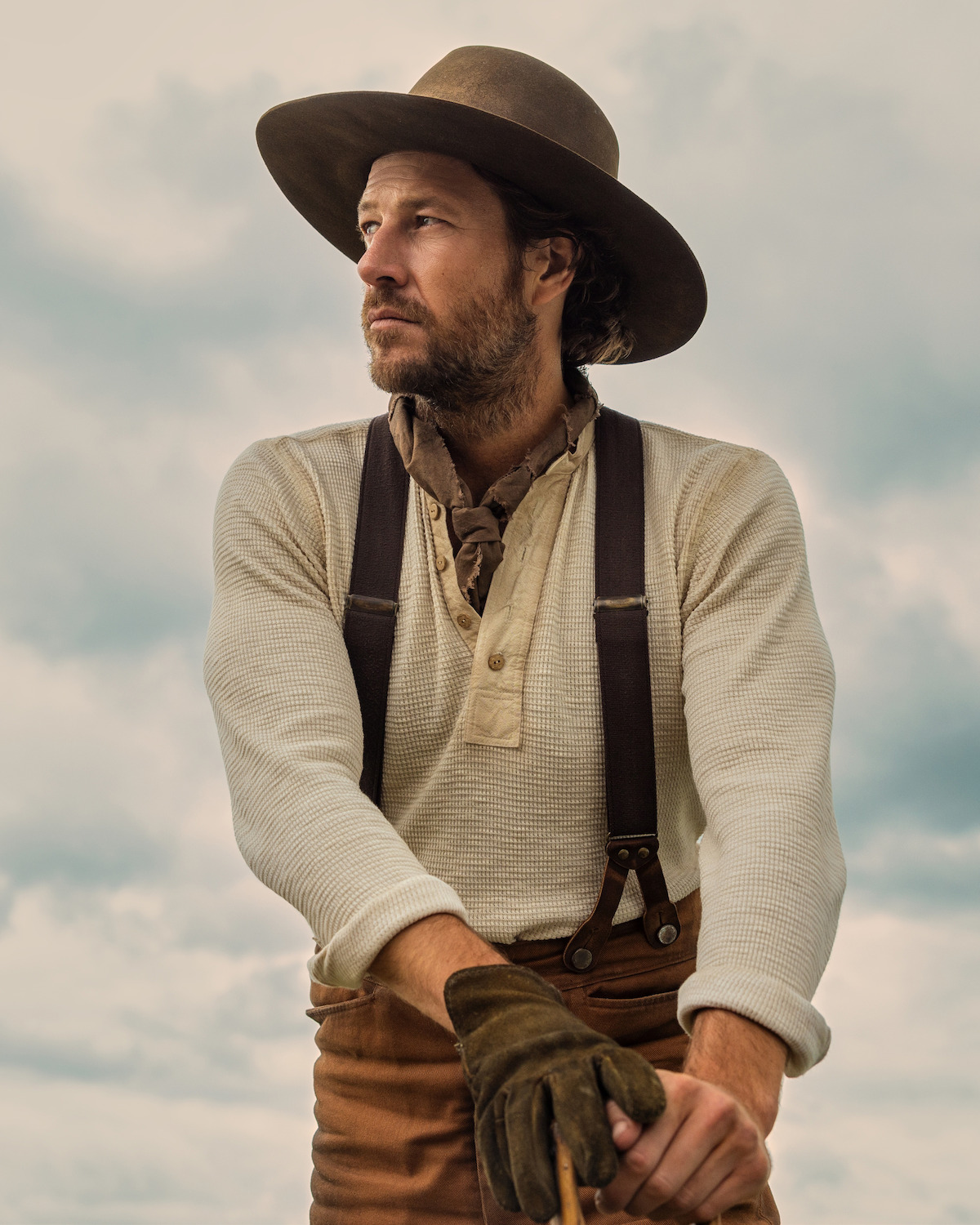 Man wearing a wide-brim hat, suspenders, gloves, and scarf stands outdoors with cloudy sky background, dressed in rustic cowboy-style clothing, gazing thoughtfully to the side.