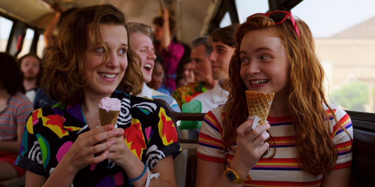 Two teenage girls enjoy ice cream cones and smile at each other on a crowded bus.