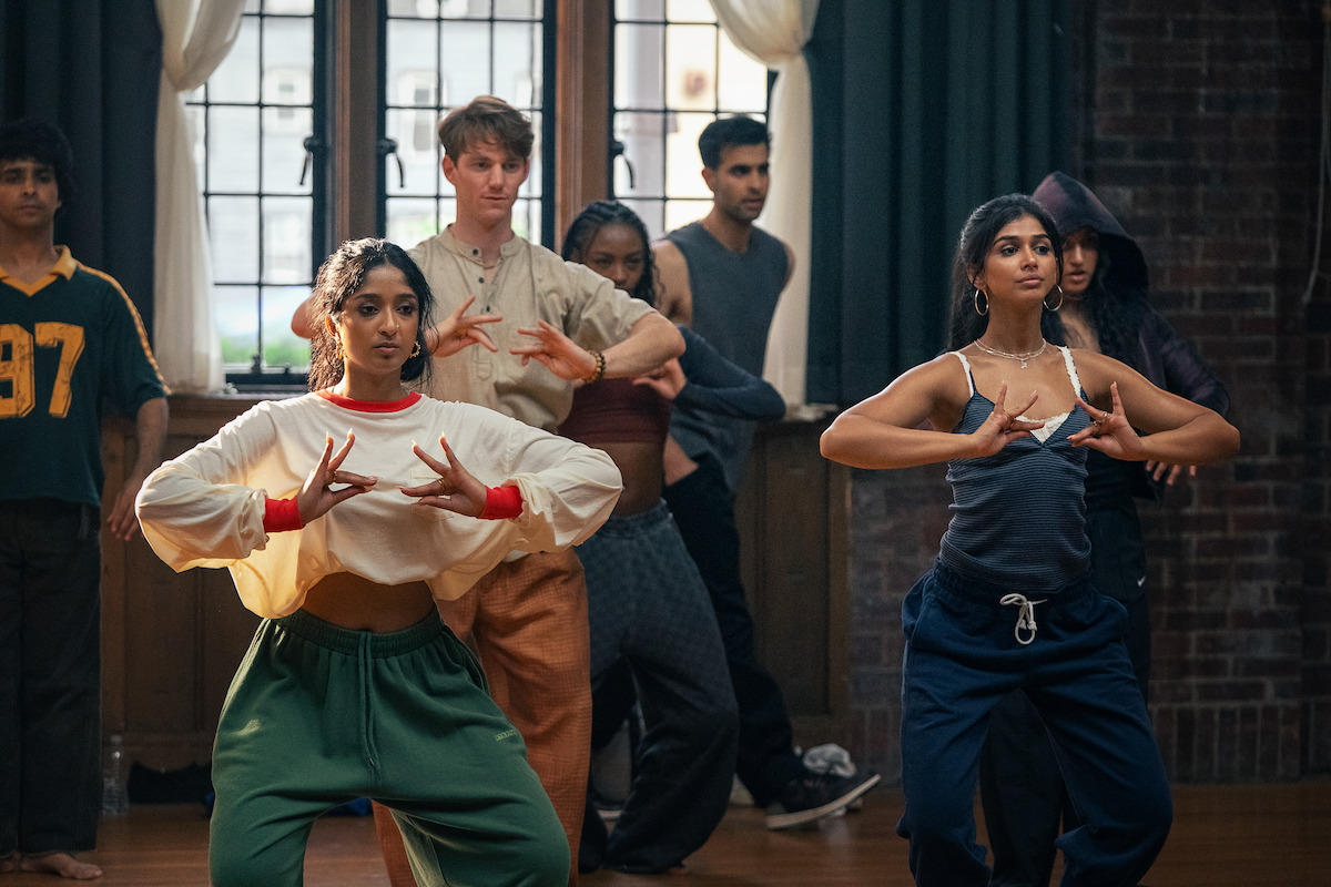 A diverse group of young adults practicing dance in a studio with wooden floors, brick walls, and large windows. The two women in the front are focused on their hand movements, suggesting a choreographed routine.