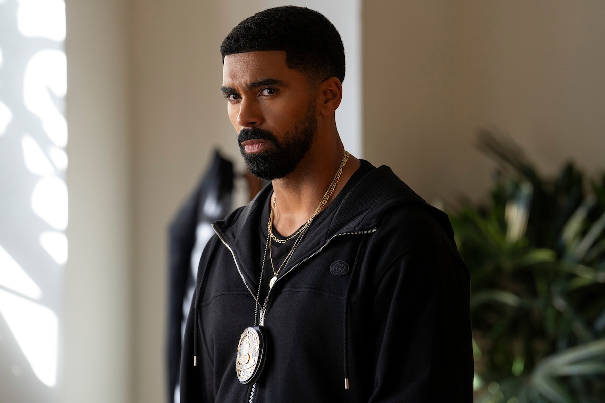 Serious man with a police badge around his neck stands indoors near plants, wearing a black hoodie, with soft light coming from the window.