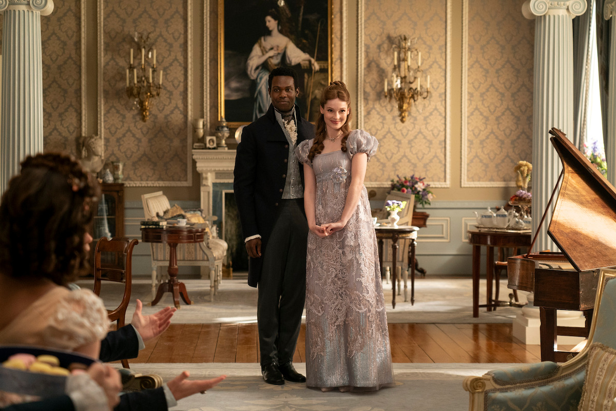 Victor Alli as John Stirling and Hannah Dodd as Francesca Bridgerton stand in a sitting room together smiling in Season 3 of ‘Bridgerton’