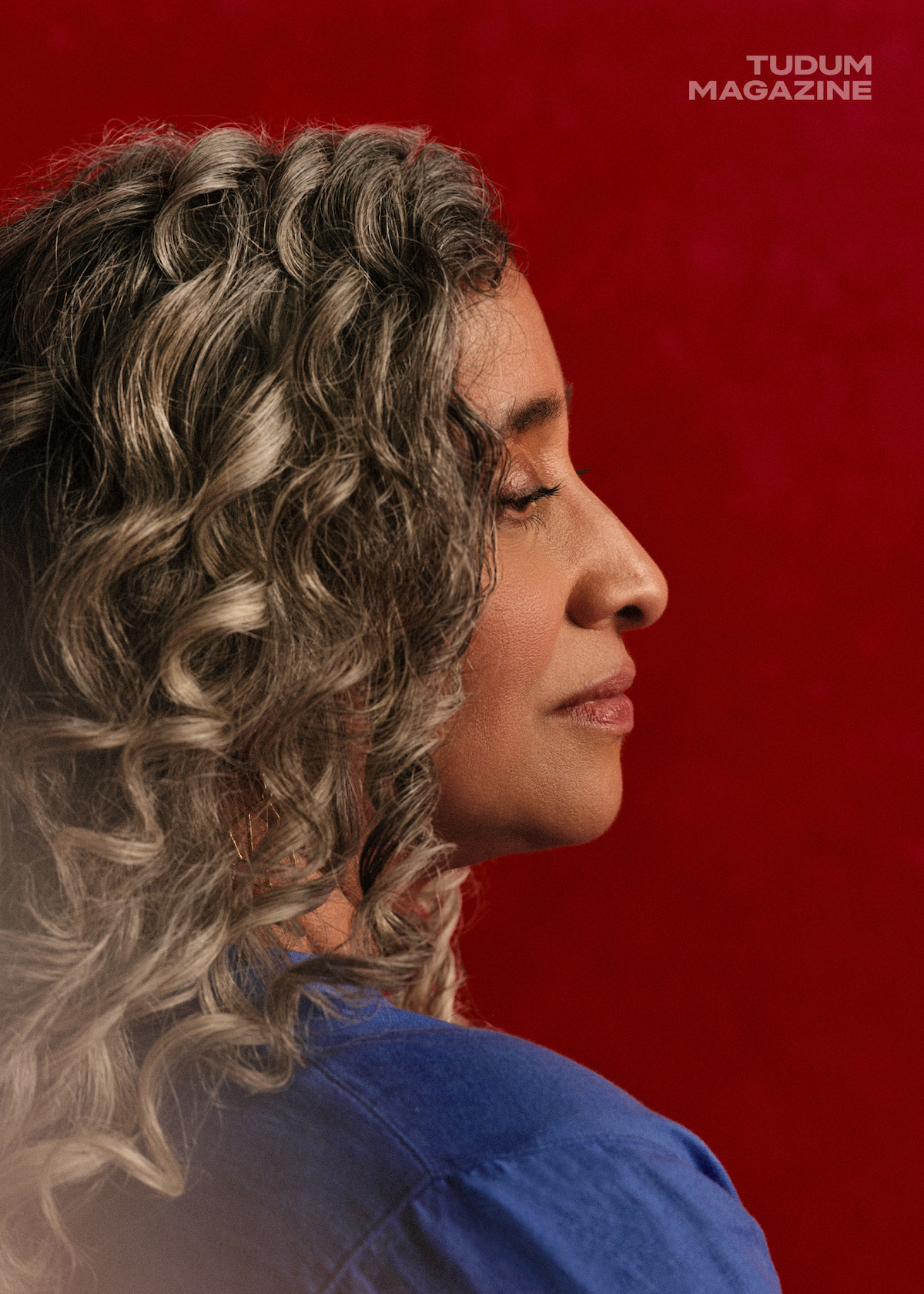 Geeta Gandbhir, director of The Perfect Neighbor, stands in front of a rich red backdrop with beautifully set curls in her graying hair, head turned away from the camera and slightly looking toward the ground with a serious expression on her face framed tightly in the crop of the image.