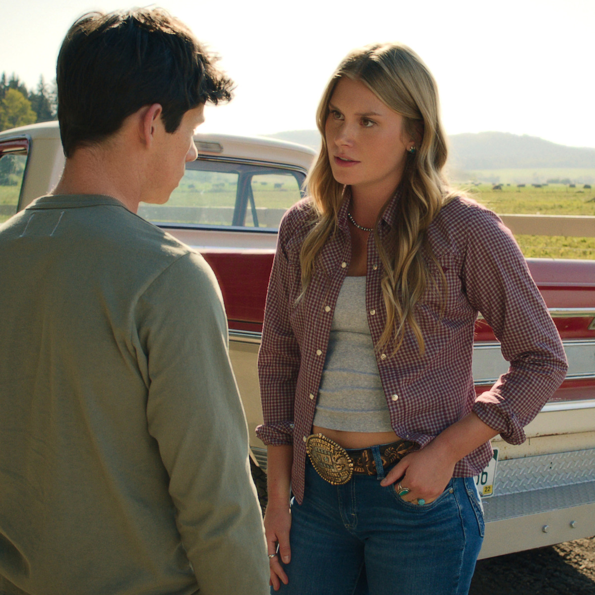 Two people are talking beside a vintage truck in a rural setting with hills and fields in the background, one wearing a casual shirt and the other in a checkered shirt with a large belt buckle.