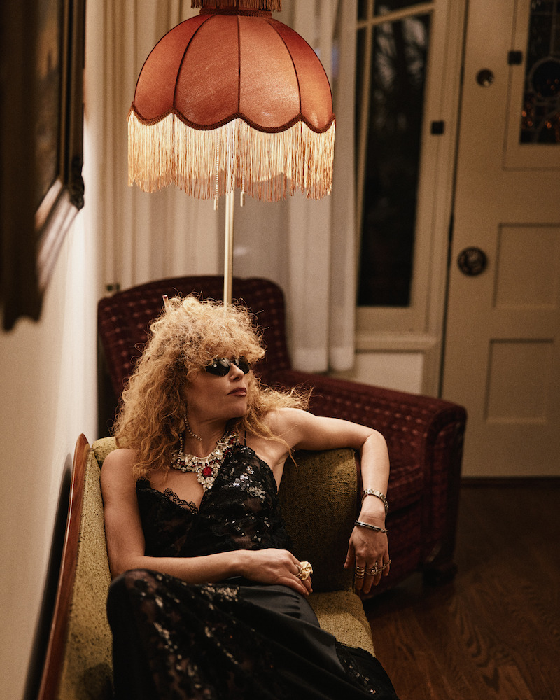 Person with curly hair and sunglasses lounges on a vintage sofa under a fringed lampshade in a dimly lit, retro-style living room with eclectic decor and warm lighting.