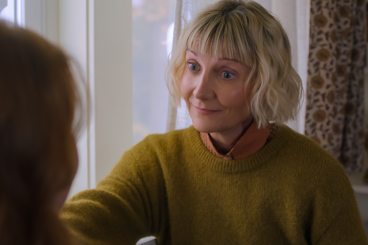 A woman with short blonde hair in a cozy mustard sweater sits indoors near a window, smiling warmly at another person with reddish hair whose back is to the camera. The room has patterned curtains and soft natural light.
