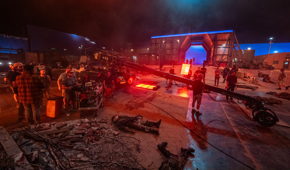 Film crew shoots a dramatic night scene on a rubble-strewn set with blue-lit futuristic gateway.