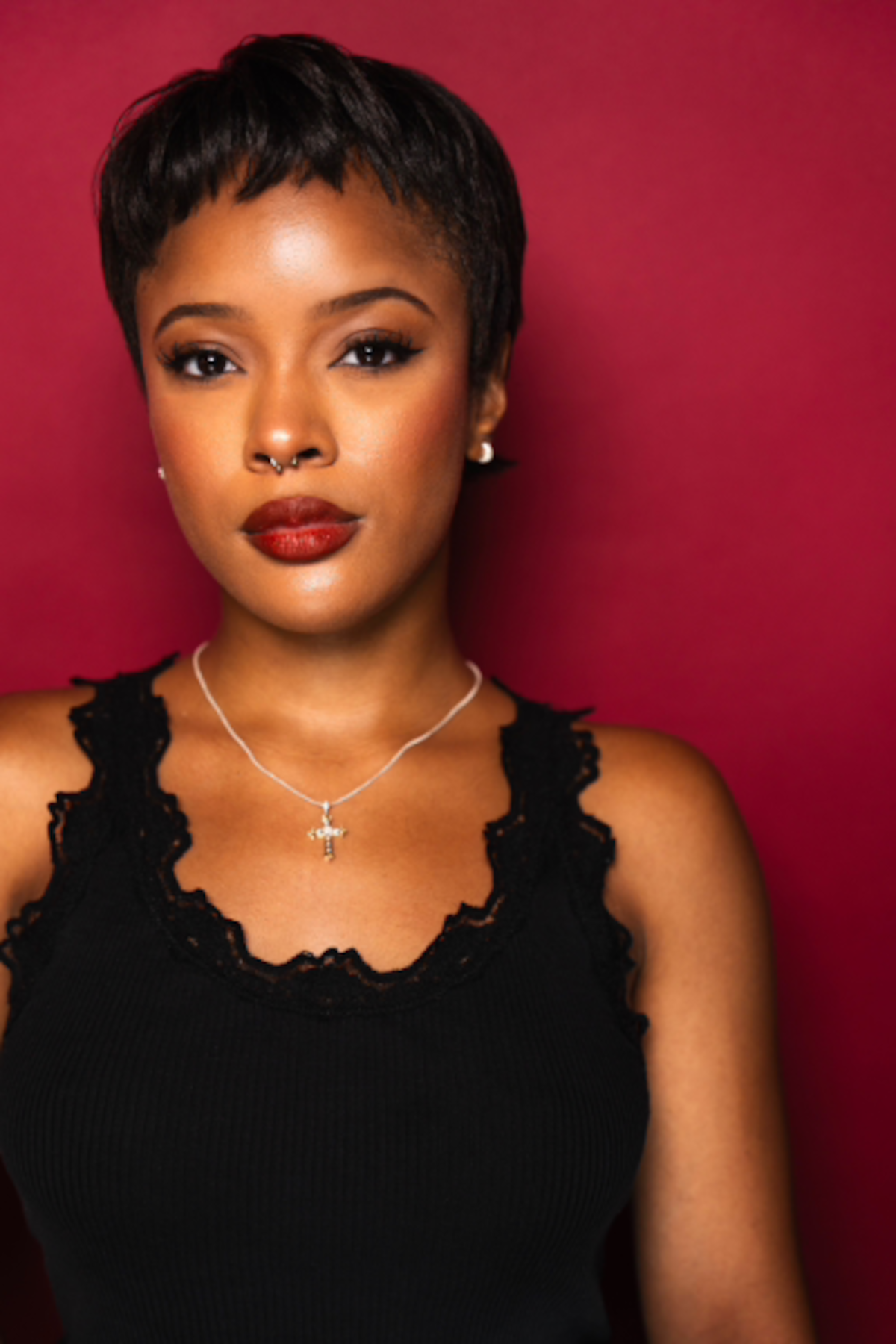 Confident woman with short hair and red lipstick, wearing a black top and silver jewelry, poses against a rich red background in a studio portrait with a bold, empowered mood.