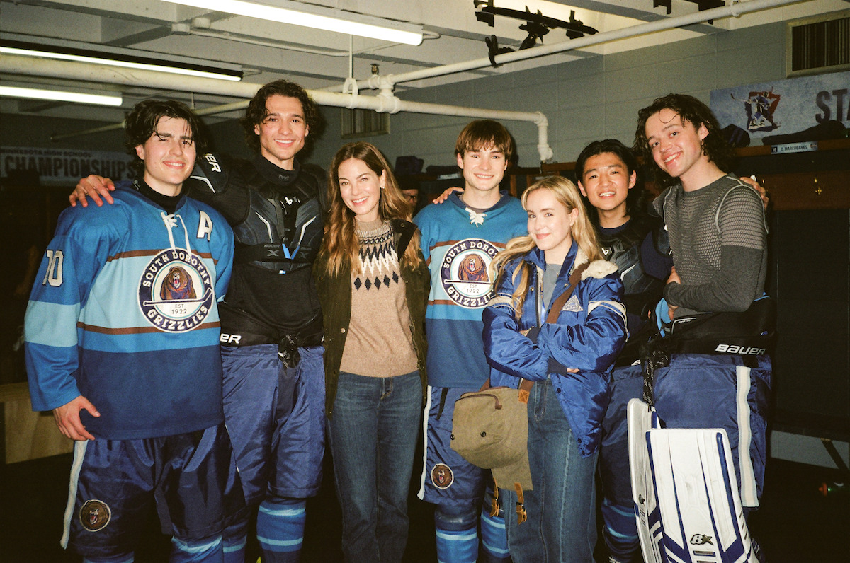 Group of young hockey players in blue uniforms and two people in casual clothes smiling together inside a locker room, with benches and hockey equipment visible in the background.