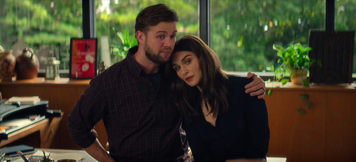 A man and woman sit close together in a cozy, sunlit office with large windows, plants, and books in the background. The man has his arm around the woman in a comforting gesture.
