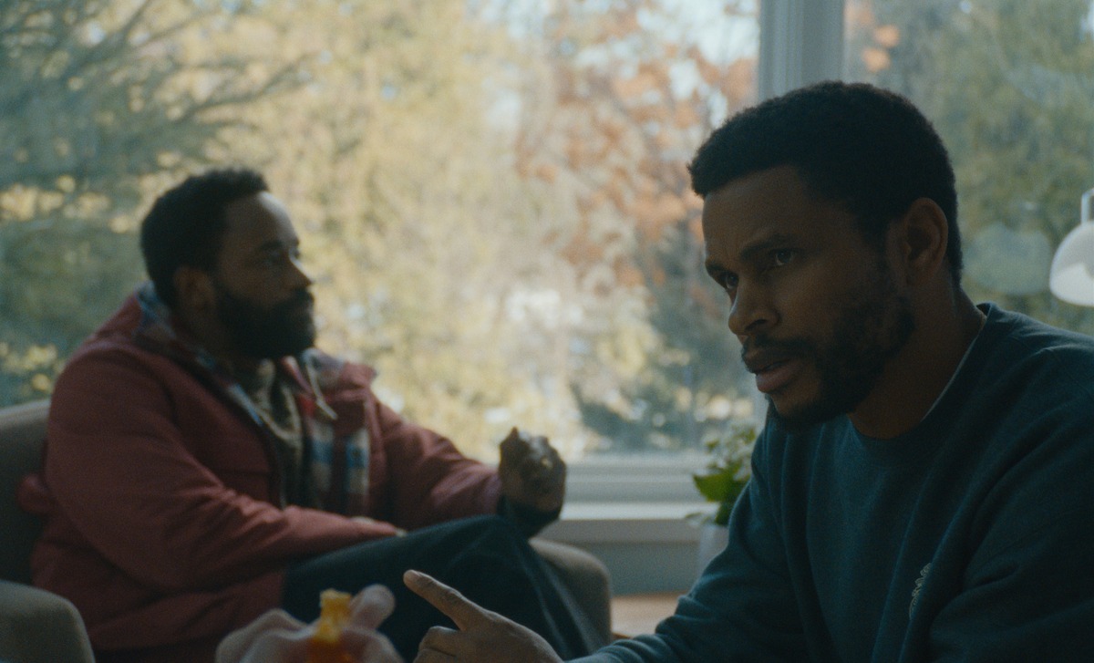 Two men sitting indoors by a large window with trees outside, engaged in a serious conversation. One man wears a red jacket with a scarf, the other a dark sweater, both in a warmly lit, calm setting.