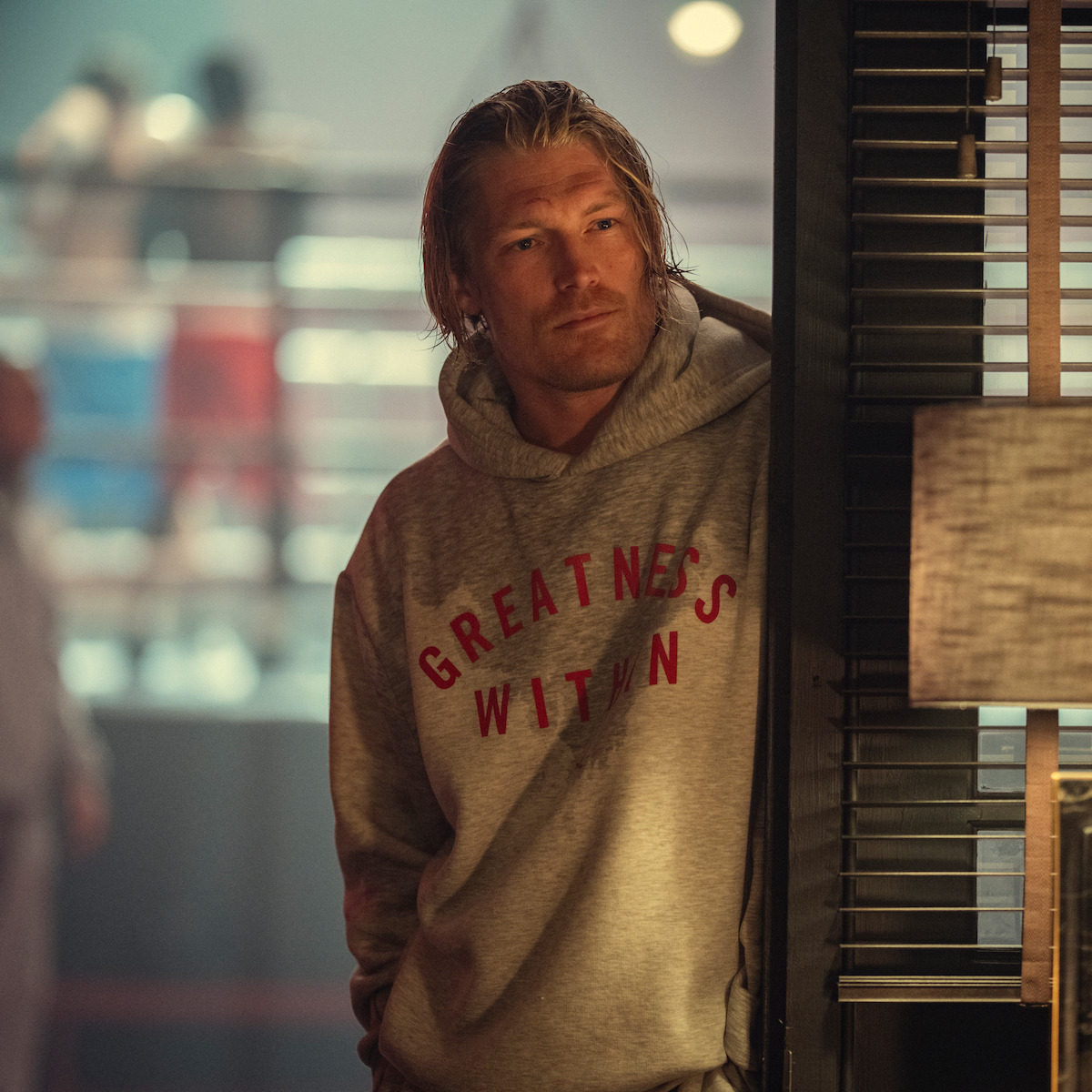 Man in a gray hoodie with "Greatness Within" text stands by a door in a dimly lit boxing gym, with fighters sparring in a ring in the blurry background.
