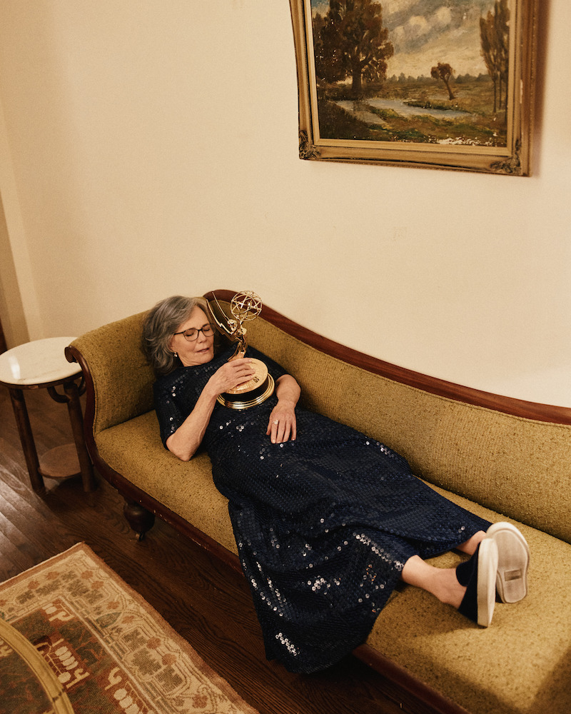 Woman in a navy sequin dress lies on a vintage couch holding an Emmy award, set in a warmly lit living room with a framed landscape painting and wooden floors.