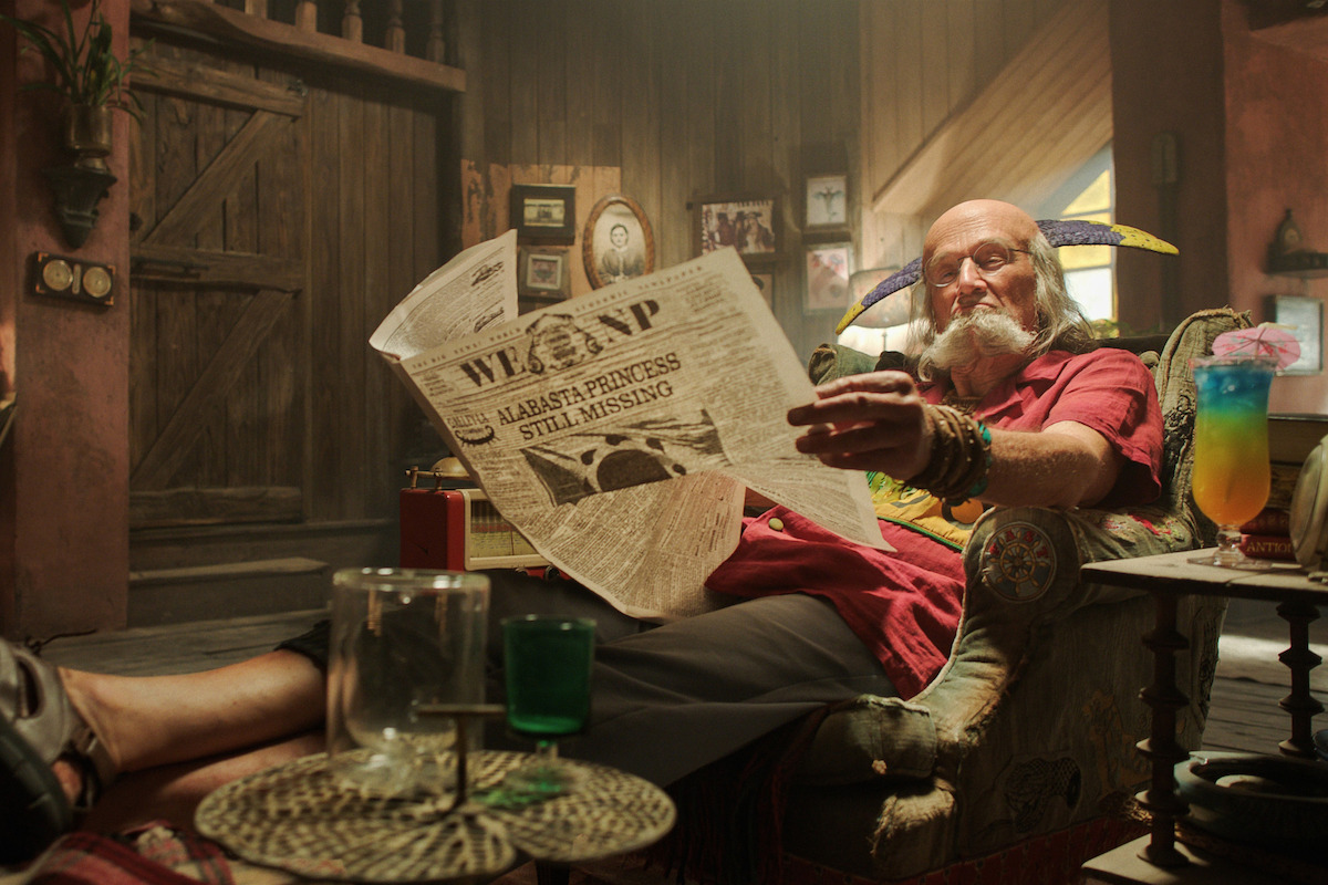 Elderly man with a jester hat relaxing in a cozy, rustic living room, reading a newspaper with "ALABASTA PRINCESS STILL MISSING" headline, surrounded by colorful drinks and eclectic decorations.