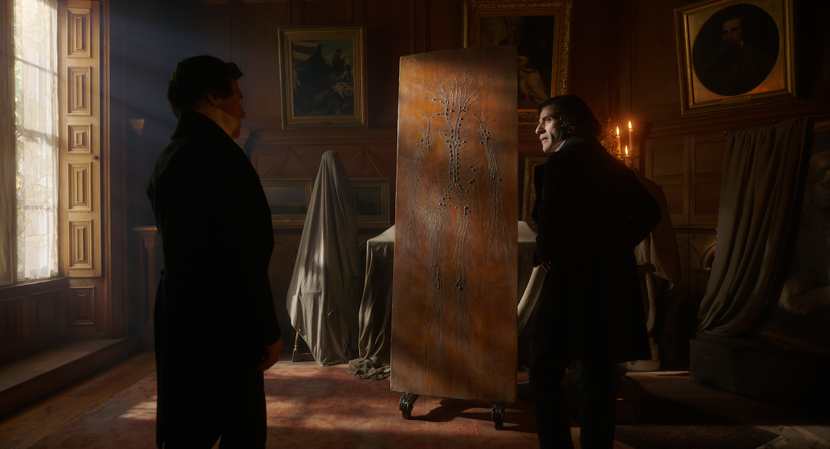 Two men in Victorian-era clothing stand in a dimly lit, wood-paneled room with framed portraits, covered furniture, and soft sunlight streaming through a window, creating a mysterious and tense atmosphere.