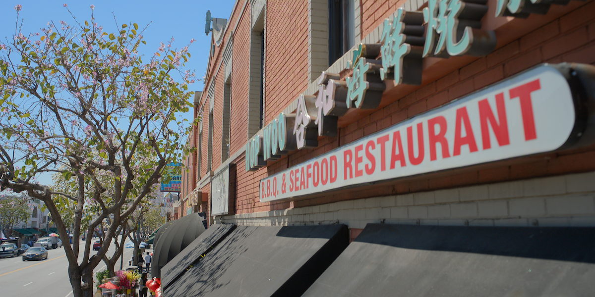 Asian BBQ and seafood restaurant exterior with red sign, brick building, and black awnings on a sunny day; cherry blossom tree and street with cars visible in the background.