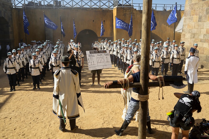 Armand Aucamp as Bogard, Aidan Scott as Helmeppo, Langley Kirkwood as Captain "Axe Hand" Morgan, and Vincent Regan as Vice Admiral Garp gather in a courtyard in Season 1 of 'One Piece.'