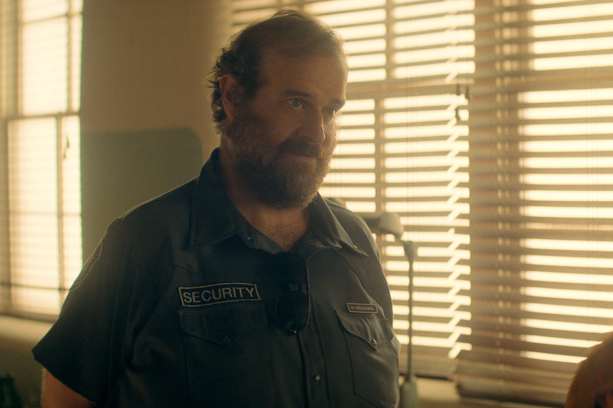 Eric Edelstein as Hank in a security uniform stands indoors beside window blinds, bathed in warm sunlight in what appears to be an office or security room.
