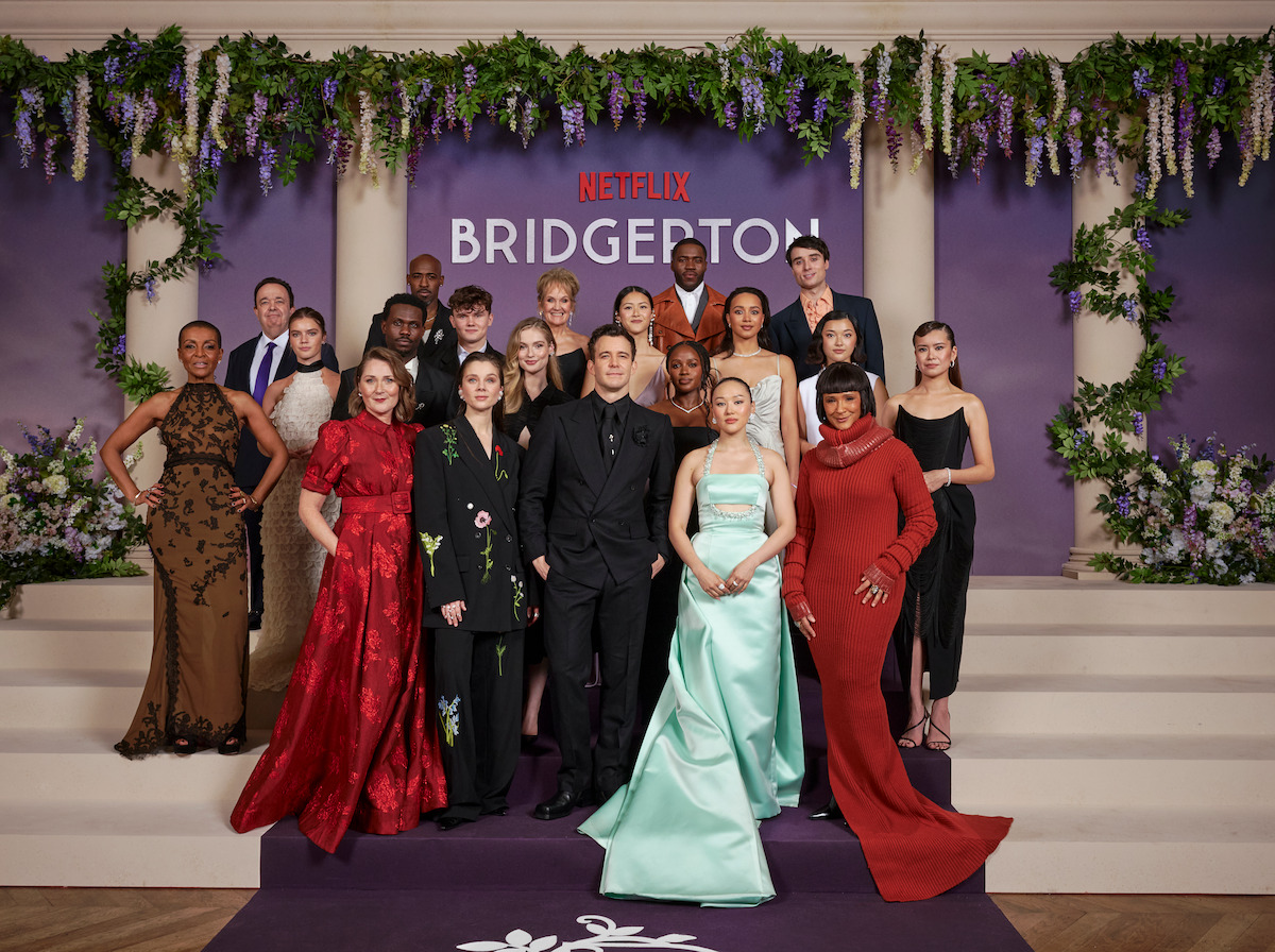 Group of people in formal attire pose on decorated purple steps at a ‘Bridgerton’ Netflix event, with floral arrangements and purple backdrop in the background.