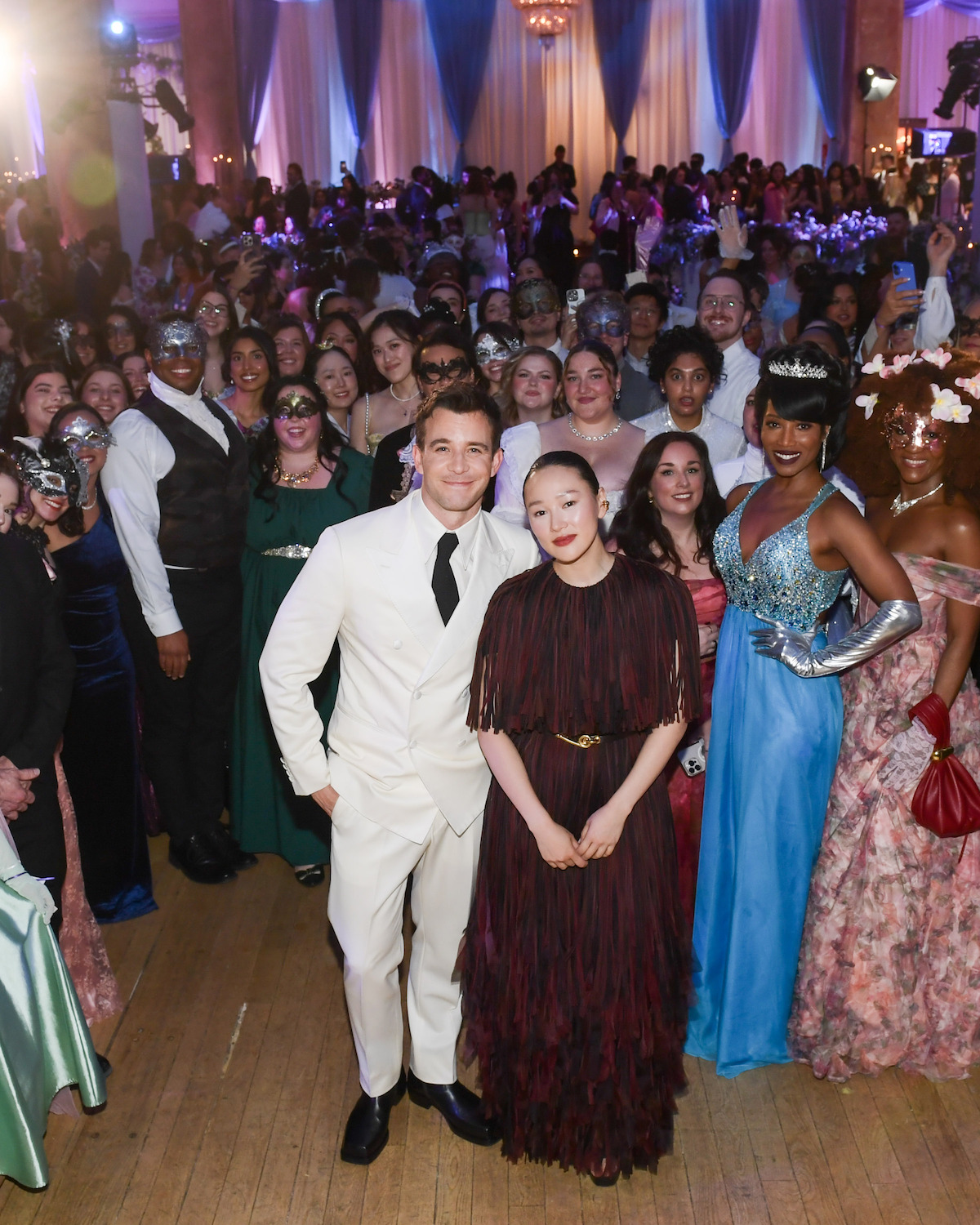 A large group of people in formal attire at a masquerade ball, with decorative dresses and suits, some wearing masks, standing in a grand ballroom decorated with drapes, chandeliers, and blue-purple lighting.